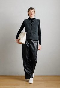 Halli tekstureeritud vest musta kõrge kaelusega jope üle, pa paired lahtiste mustade nahkpükstega ja valgete tossudega, hoides kreemikat kotikest.