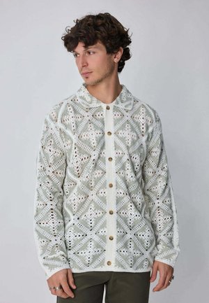 Homme aux cheveux bouclés portant un cardigan en crochet beige et blanc à motif, avec des boutons marron, regardant sur le côté devant un fond uni.