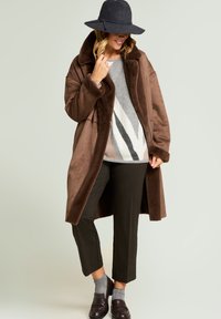 Brązowy, oversize płaszcz z kołnierzem z futra syntetycznego, szary sweter w wzory, ciemne spodnie, szare skarpetki i ciemne buty. Modelka nosi czarny filcowy kapelusz.