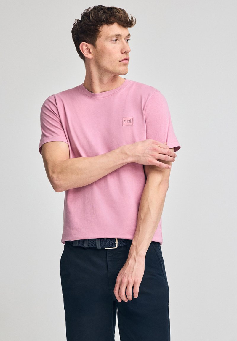 T-shirt di cotone rosa chiaro con colletto tondo e maniche corte, caratterizzata da un piccolo logo quadrato sul lato sinistro del petto. Indossata con pantaloni blu navy.