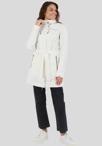 Witte waterdichte jas met een tailleband, ritszakken en verstelbare capuchon. Draagt met zwarte broek en witte platform sneakers.