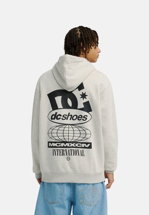 Osoba z krótkimi twistami nosi jasnoszarą bluzę z kapturem z dużym logo DC Shoes i grafiką globusa, zestawioną z jasnoniebieskimi dżinsami.