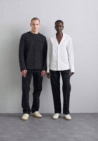 Two individuals stand side by side. One wears a textured black sweater and dark pants; the other, a white button-up shirt and dark cargo pants.