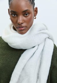 Bijela, pahuljasta šal obavijen oko vrata osobe, s mekim teksturom i suptilnim prugastim uzorkom, u kombinaciji s tamnozelenim puloverom.