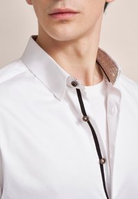 Weiße Button-up-Hemd mit einem kontrastierenden braunen gemusterten Kragen, schwarz abgesetzt und runden Knöpfen entlang der Knopfleiste, glatter Stofftextur.