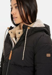 Zwarte pufferjack met een getextureerde fleece voering, contrasterende beige kraag, ritssluiting aan de voorkant en decoratieve koorden met plastic sluitingen.
