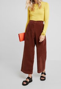 Žlutý dlouhý rukáv a hnědé široké kalhoty z manšestru. Černé sandály s dvojitým páskem. Oranžové psaníčko přidává kontrastní detail.