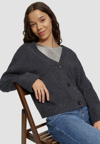 Dunkelgraue gerippte Strickjacke mit V-Ausschnitt, drei schwarzen Knöpfen und langen Ärmeln, getragen über einem silbernen Oberteil und kombiniert mit blauen Jeans.
