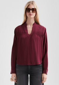 Burgunderfarbene Bluse mit langen Ärmeln, einem Ausschnitt mit Schlitz, gelegten Details auf der Vorderseite und taillierten Manschetten. Getragen mit dunklen Jeans und Sonnenbrille.