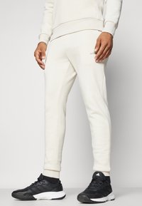 Light beige joggers with a tapered fit, made of soft fabric; featuring side pockets and a subtle logo near the right pocket. Paired with black sneakers.