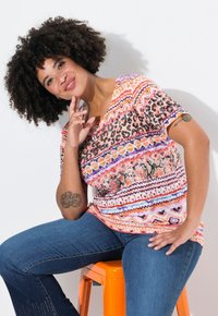 Multifarvet mønstrede t-shirt med orange, lilla og sorte designs, parret med blå jeans. Sidder på en orange skammel.