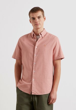 Giovane uomo con indosso una camicia rosa a maniche corte sopra una t-shirt bianca e pantaloni verdi, in piedi davanti a uno sfondo neutro.