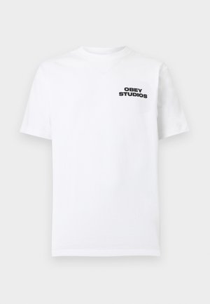 Fehér, rövid ujjú, kerek nyakú póló fekete "OBEY STUDIOS" felirattal a bal felső mellrészen, sima háttér előtt.