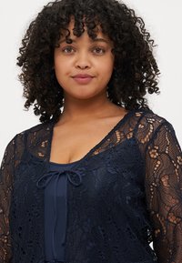 Cardigan in pizzo navy, maniche lunghe, presenta motivi floreali, apertura frontale con chiusura a fiocco, indossato sopra un top navy a tinta unita. Texture liscia.