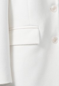 Blazer blanc en tissu texturé, avec une poche unique et trois boutons crème légèrement brillants. Lignes épurées et coupe ajustée.