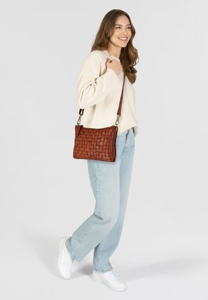 Bolso de cuero trenzado en marrón, de forma rectangular compacta, con una correa larga y cierre con cremallera. Combinado con unos vaqueros claros y un suéter de color crema.