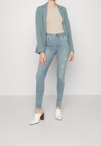 Jeans de mezclilla de tiro alto en azul claro con detalles desgastados, combinados con una camisa ajustada color beige y un blazer en tono azul verdoso. Mule blancas con tacón completan el look.