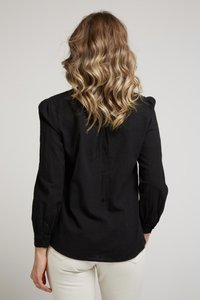 Chemise noire à manches longues en tissu texturé, avec des épaules bouffantes, des poignets boutonnés et un ourlet arrondi, portée avec un pantalon de couleur claire.