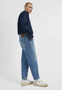 Jeans azzurri chiari con una vestibilità rilassata, caratterizzati da una patta in pelle marrone sul retro, abbinati a una camicia in denim blu scuro e a scarpe da ginnastica bianche.