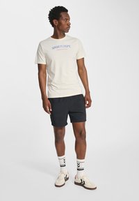 Lysbeige t-shirt med blå tekst "SPORTS DEPT." parret med sorte shorts, hvide sokker med sorte striber og beige sneakers med sorte detaljer.