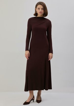 Ung kvinde stående iført en langærmet, gulvlængde mørkebrun kjole og matchende kitten heel-sko mod en ensfarvet baggrund.