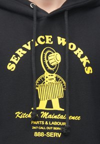 Čierna mikina s kapucňou so žltou grafikou zobrazujúcou kľúč a textom "SERVICE WORKS" a "Kitchen Maintenance" s nastaviteľnými šnúrkami na krku.