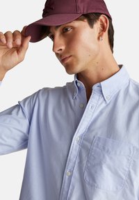 Světle modrá košile na knoflíky s kapsou na hrudi, vyrobená z hladké látky. Spárována s vínovou čepicí s vyvýšeným logem.