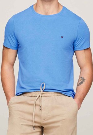 Mand iført en tætsiddende lyseblå kortærmet t-shirt og beige bukser med snor, med hænderne i lommerne.