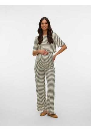 Femme souriante portant une tenue deux pièces rayée noir et blanc, debout avec une main sur le ventre et l’autre sur la hanche, chaussée de ballerines beige.