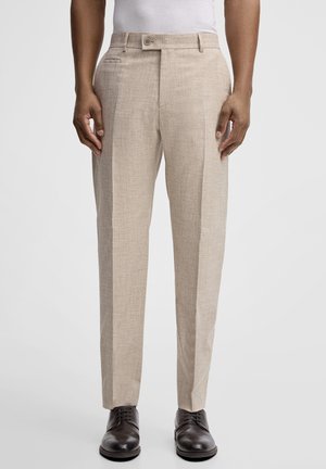 Mann trägt beige, maßgeschneiderte Hosen mit Bügelfalte vorne, weißes Hemd, in die Hose gesteckt, und dunkelbraune Lederschuhe, steht vor einfachem Hintergrund.