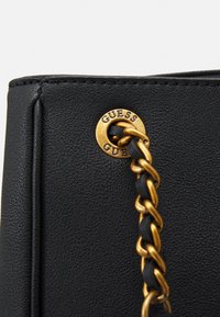 Mustan nahkainen käsilaukku, jossa on kultasävyiset ketjuhardwaret ja brändätty metallilappu, jossa on kohokuvioitu "GUESS". Huomioi teksturoitu pinta.