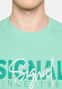 Camiseta verde menta con cuello redondo, que presenta un gran gráfico de "SIGNAL" en texto en cursiva de color verde oscuro y blanco. Tejido suave y liso.