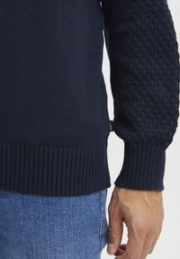 Tengerészkék kötött pulóver ribbelt mandzsettával és alsó résszel; texturált minta a ujjakon; kék farmernadrággal párosítva, bemutatva egy laza stílust.