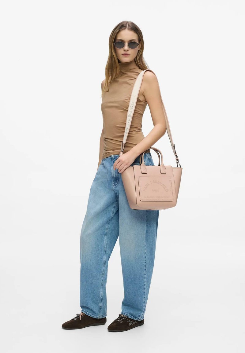 Femme portant des lunettes de soleil rondes, un top marron sans manches, un jean bleu large et des chaussures foncées, tenant un sac à main beige avec une bandoulière.
