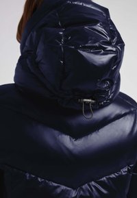 Marineblå dunjakke med høj krave og indsnøring i hætten. Skinnende, glat tekstur med chevron-syningsdetaljer.