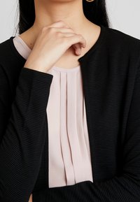 Czarny, prążkowany kardigan z okrągłym dekoltem, nałożony na jasnoróżową bluzkę z plisami. Gładka tekstura z widocznymi detalami tkaniny.