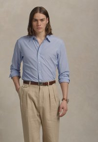 Camisa de mangas longas azul clara combinada com calças beje plissadas e um cinto castanho. Tecido suave com colarinho abotoado e detalhe de relógio.