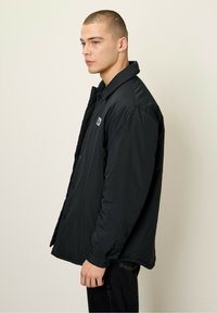 Black button-up jacket made of lightweight fabric, featuring a soft texture, relaxed fit, and a small logo on the left chest area.