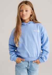 Sweat à capuche bleu clair avec une poche kangourou, des poignets et un ourlet côtelés, arborant un logo à l'avant. Porté avec un jean en denim clair. Texture douce.