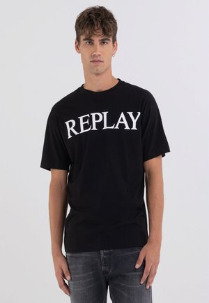 Camiseta de algodón negra con un gran estampado blanco de "REPLAY" en la parte frontal, mangas cortas y un corte relajado, combinada con jeans de mezclilla grises.