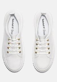 Baskets blanches à enfiler en toile avec œillets en métal doré, surface texturée, col rembourré et semelles en caoutchouc blanc. Logo Timberland visible à l'intérieur.