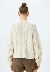 Sweterek w kolorze kremowym z dzianiny, z wzorami w warkocze i fakturowanymi frędzlami na rękawach. Ribbowe wykończenie. Krótkie szorty we wzór jodełki.