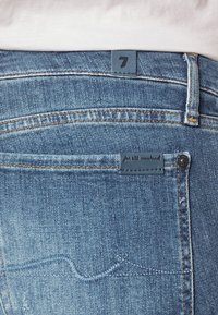 Vaqueros de denim con un lavado azul claro, con una parche de cuero en la parte posterior etiquetado como "7" y un diseño sutil en el bolsillo trasero con acentos de costura amarilla.
