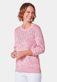 Hellrosa gestrickter Pullover mit einem strukturierten Spitzenmuster auf der Vorderseite und Dreiviertelärmel. Gerippter Saum und Bündchen. Kombiniert mit weißen Hosen.