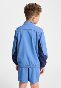 Ljusblå sportjacka med marinblå ärmar och reflexdetaljer. Tyget verkar slätt med ett elastiskt midjeband, åtföljt av matchande shorts.