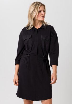 Zwarte shirtjurk met lange mouwen, twee borstzakken, een trekkoord in de taille en subtiele studs. Zacht materiaal en een ontspannen pasvorm.