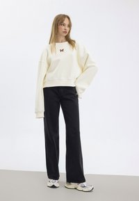 Felpa color crema con maniche oversize, decorata con un ricamo di farfalla. Abbinata a jeans neri a vita alta e a gamba larga, insieme a scarpe da ginnastica.