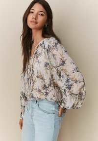 Bluză florală ușoară și transparentă cu imprimeuri botanice galbene și albastre, mâneci elasticizate și un decolteu cu șnur, asortată cu blugi bleu deschis.