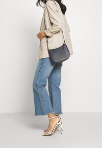 Námořnická crossbody kabelka s hladkou texturou a zlatými detaily, nošena s béžovým blazerem a světle modrými zvonovými džíny a bílými řemínkovými podpatky.