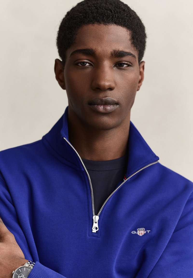 Young man with short curly hair wearing a blue quarter-zip sweatshirt and a silver wristwatch, facing forward with a neutral expression.